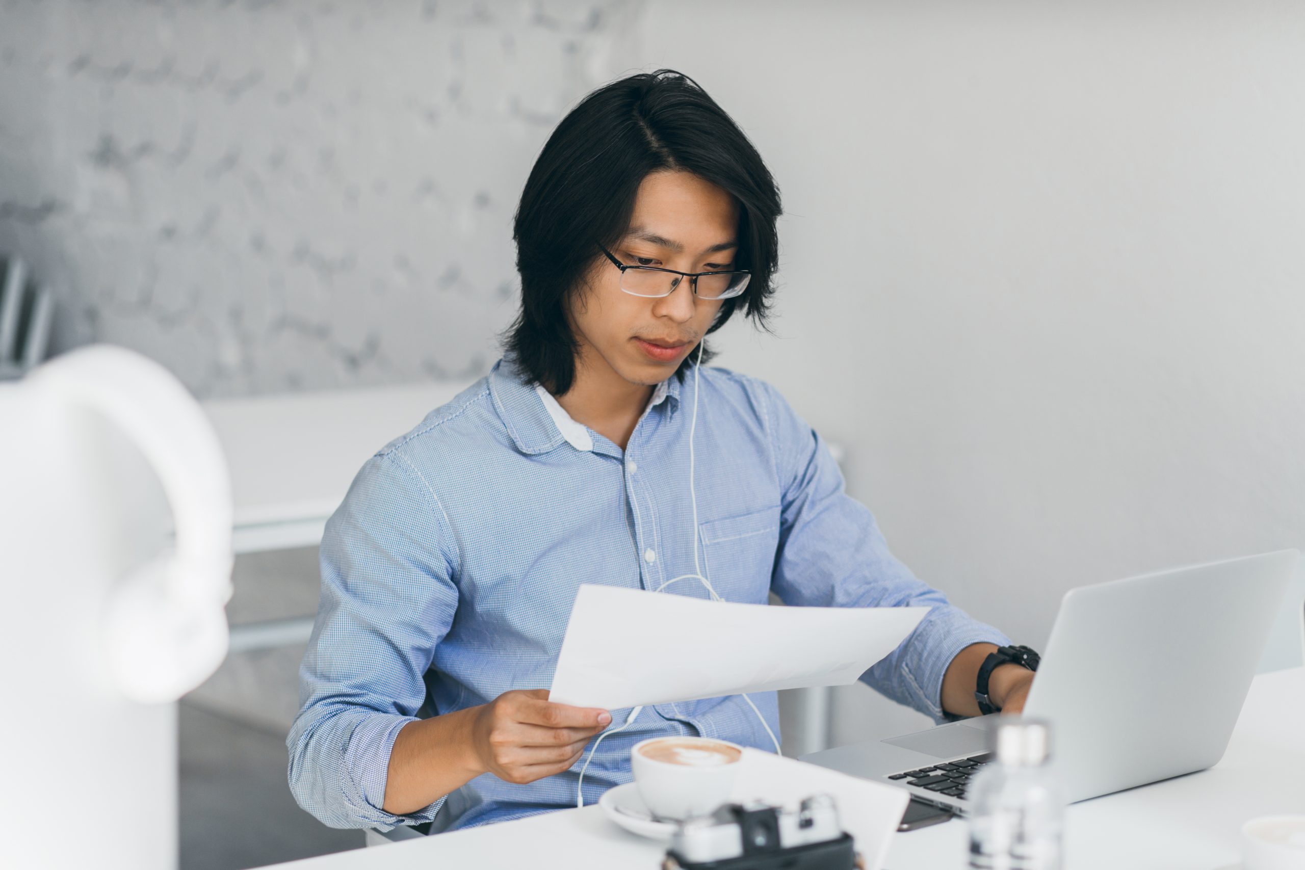 concentrated asian office worker in earphones reading documents at workplace. indoor portrait of chinese freelance it specialist drinks coffee while using with laptop..